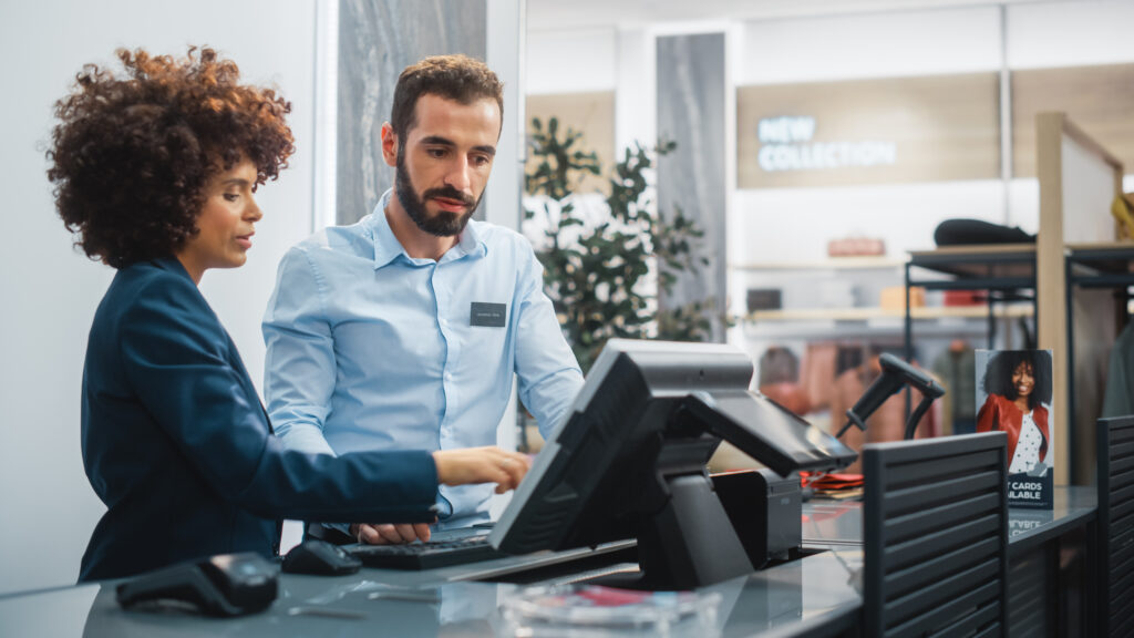 Clothing Store Checkout Cashier Counter: Female Retail Sales Manager Explaining to New Male Assistant Employee Everything He Needs to Know About Working at Fashionable Shop with Stylish Brand Designs.